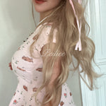 Woman with long blonde hair wearing a pink floral dress, standing against a light-colored wall.