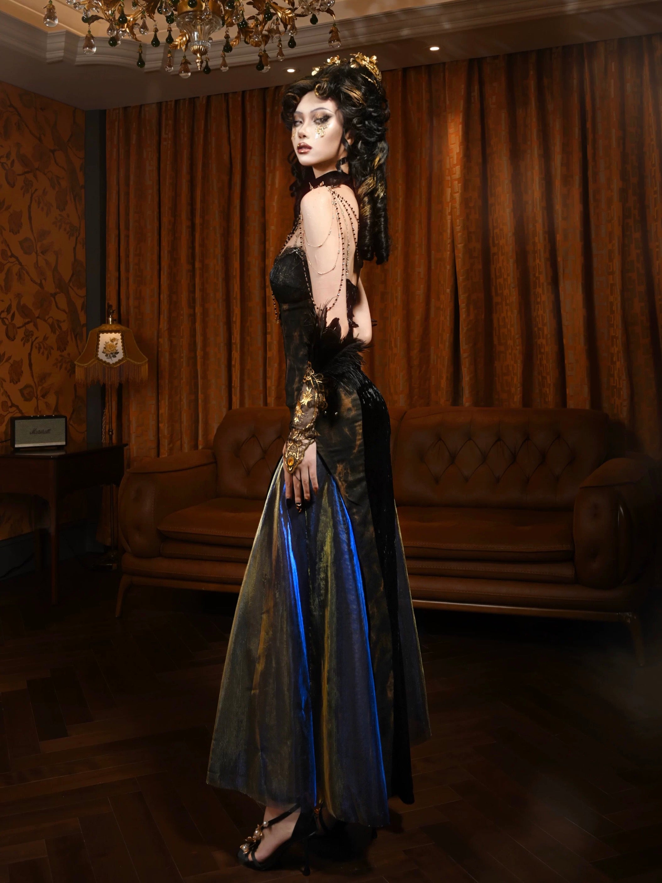 Woman in a long, dark dress with blue accents standing in an elegant room with brown curtains and a chandelier.