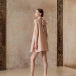 Woman in a light pink dress with a bow standing against a textured wall.