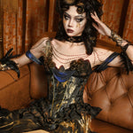 Woman in a dark, ornate Blood Supply Golden Hour Baroque Corset with gold accents sitting on a brown couch.