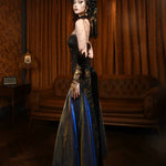 Woman in a long, dark dress with blue accents standing in an elegant room with brown curtains and a chandelier.