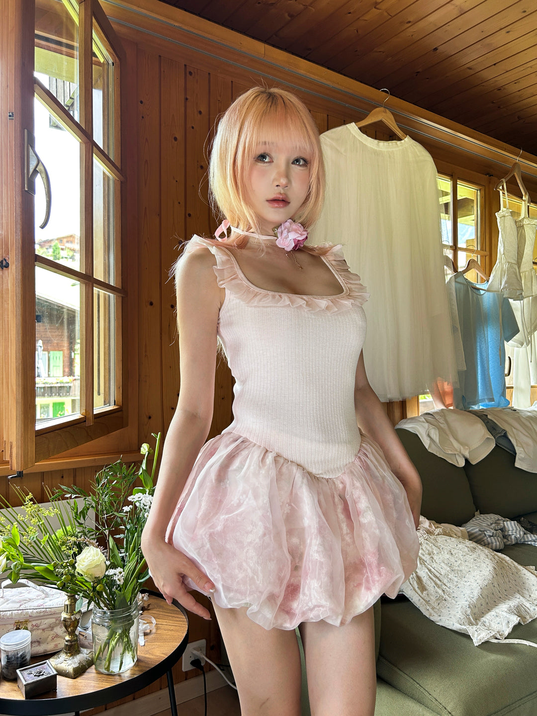 Teenager girl in pink hair wearing a pink dress standing in a room with wooden walls and a window.