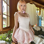 Teenager girl in pink hair wearing a pink dress standing in a room with wooden walls and a window.
